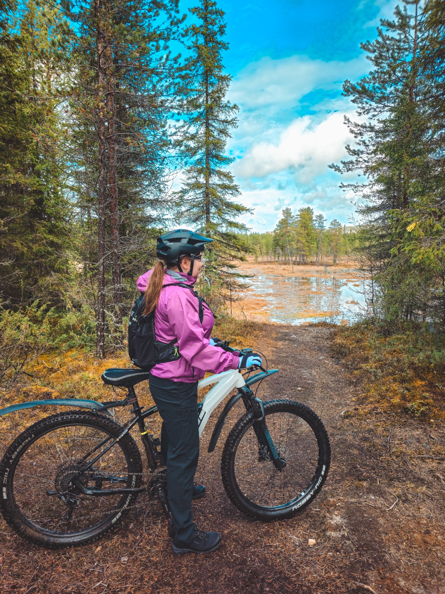 Pyöräilijä seisoo maastopyörän vieressä metsäpolulla, sininen taivas ja lammikko näkyvät taustalla.
